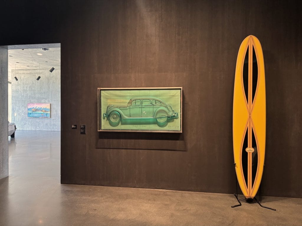 A translucent green sculpture of a vintage car in a glass case hangs next to a tall, yellow surfboard inside a dark-walled museum gallery.