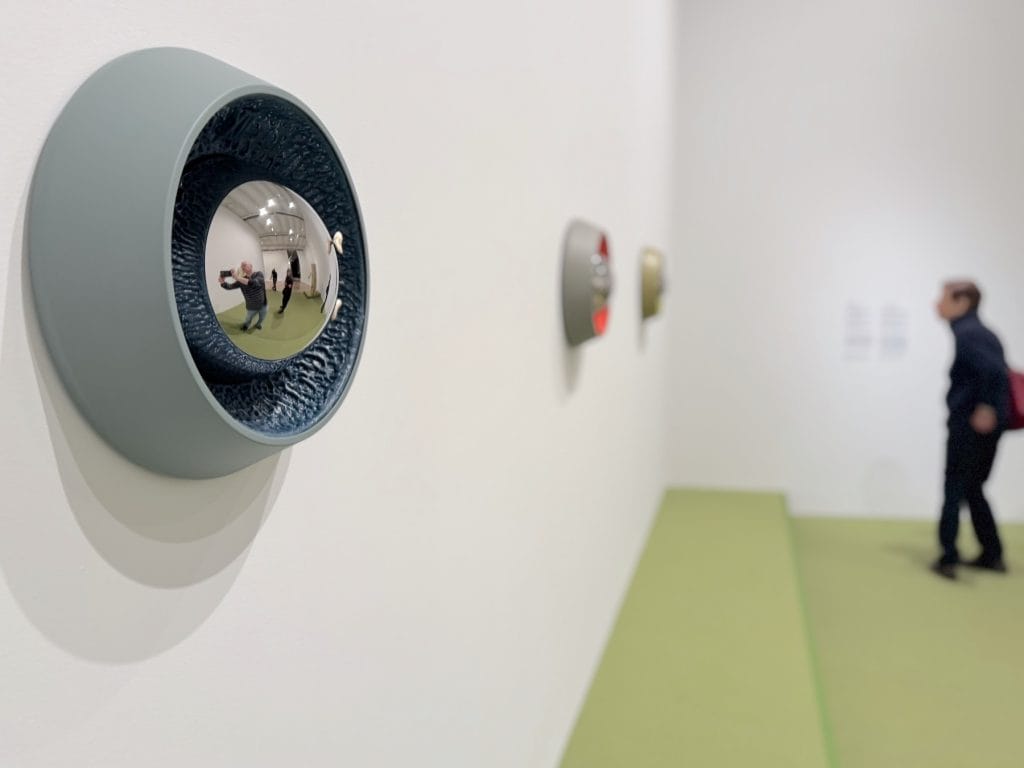 An installation view of Cooper Jacoby’s sculptures at the Whitney Biennial, showing three circular wall-mounted works with reflective convex centers along a white gallery wall.