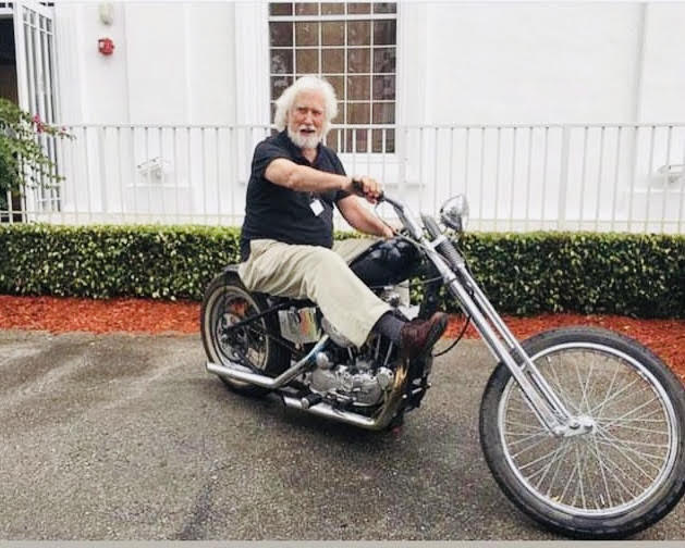 a white haired man sits on a motorcycle inthe middle of a street