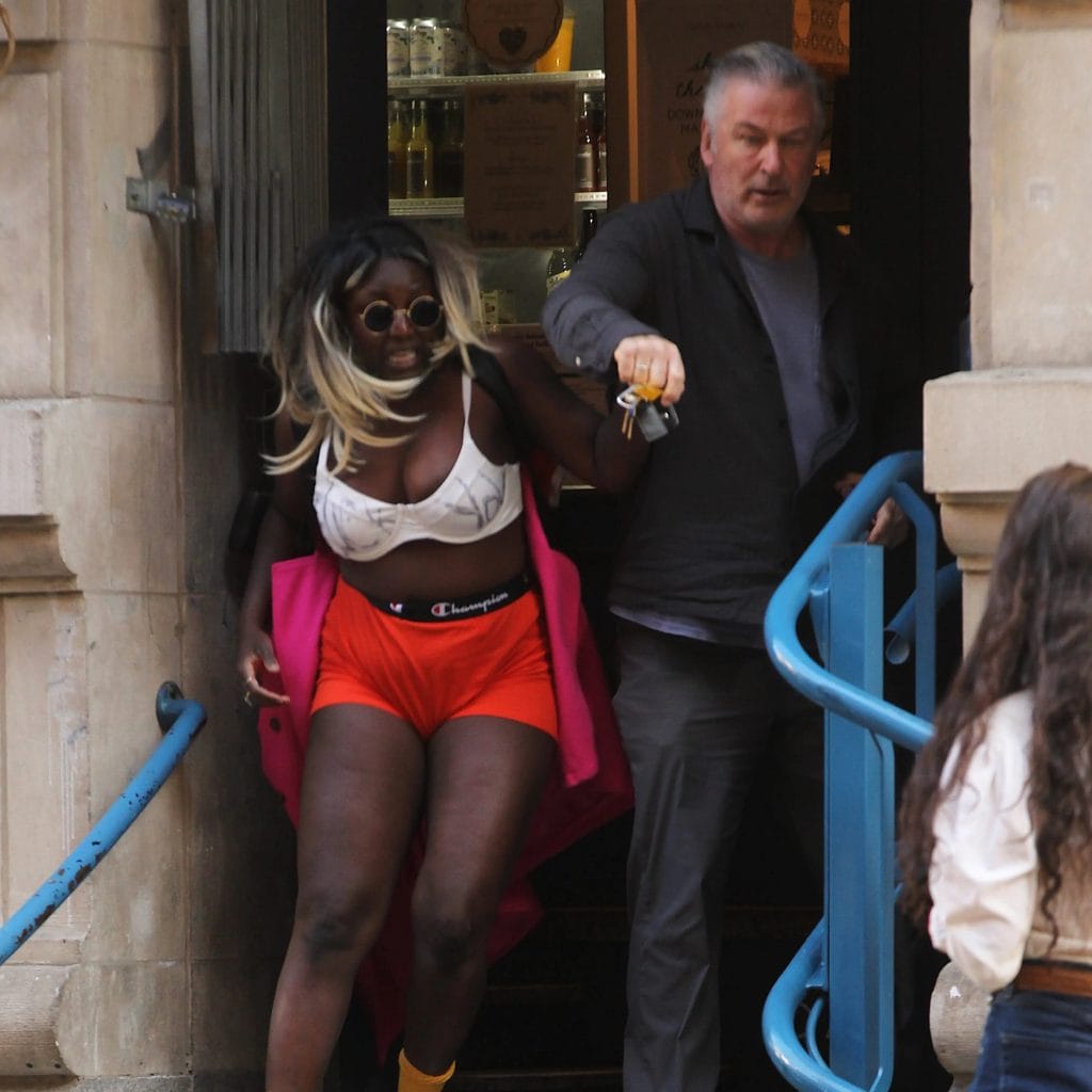 A photograph of a skimpily dressed Black women rushing out of an establishment with Alec Baldwin looking angry behind her