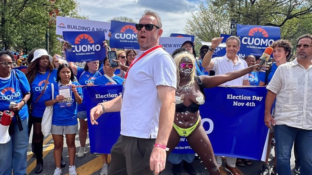 A photograph of a barely-dressed Crackhead Barney standing with her arms out in front of a procession by Andrew Cuomo and his supporters bearing a banner, with a tall man in sunglassses standing in front of her