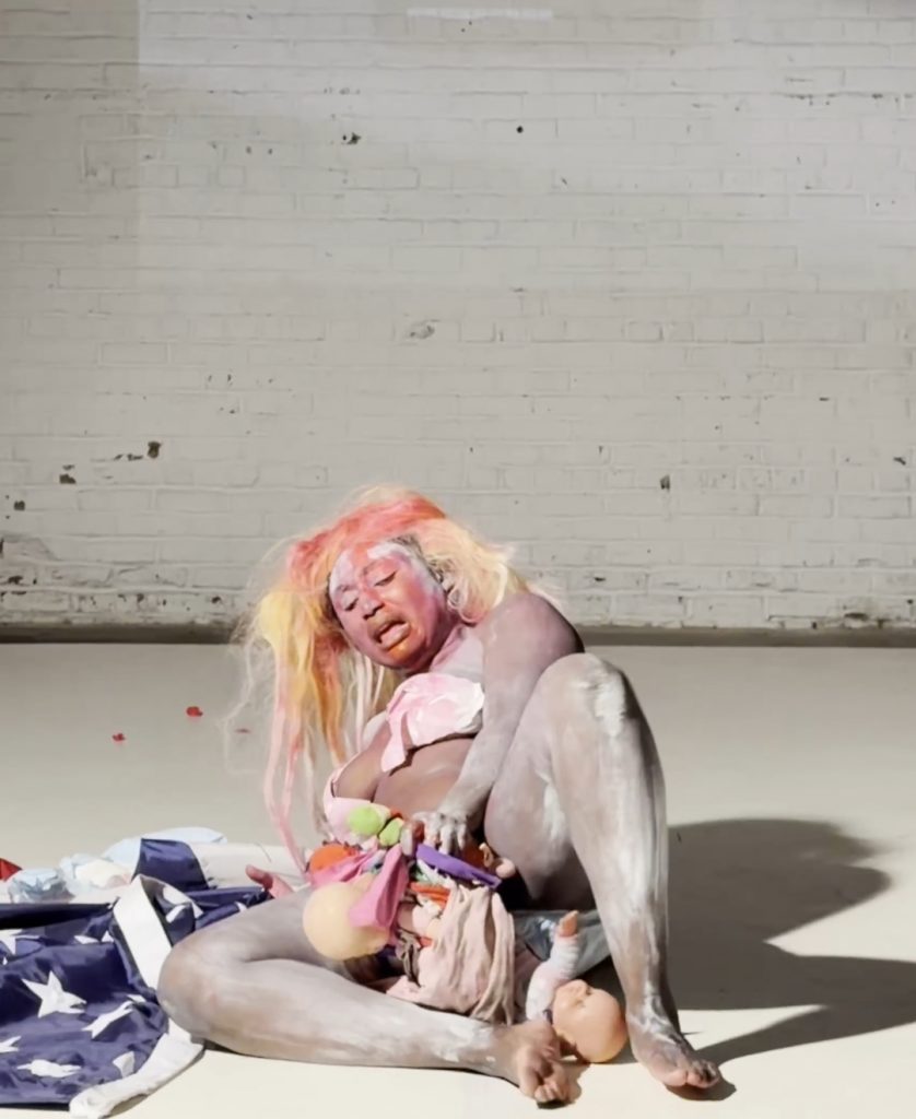 A photograph of a Black woman painted white laying on the floor almost nude giving birth to a plethora of dolls on a white theater stage