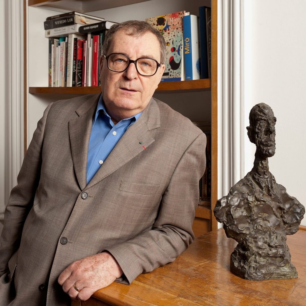A photo of an older white man with short graying hair and glasses, wearing a light brown suit and blue shirt, leaning against a wooden table. Beside him sits a rough-textured bronze bust sculpture. A bookshelf filled with art books is visible behind him.