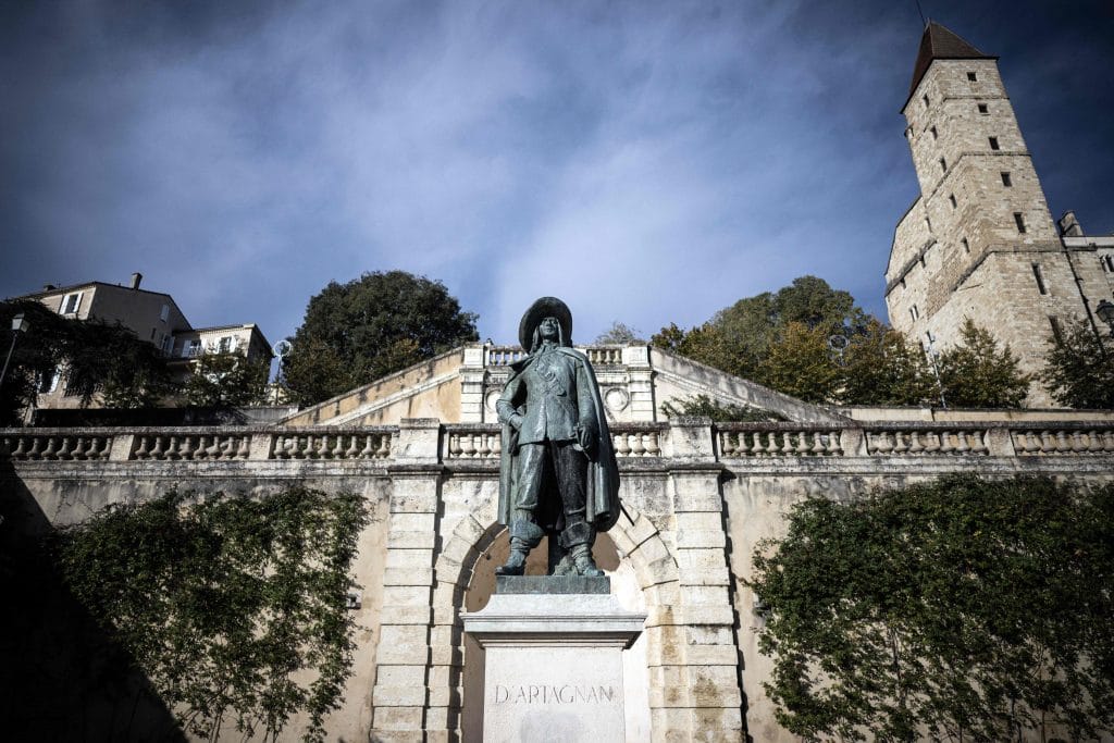 A photograph of a sculpture depicting D'Artagnan in front of a white stone structure, beneath a blue and slightly cloudy sky