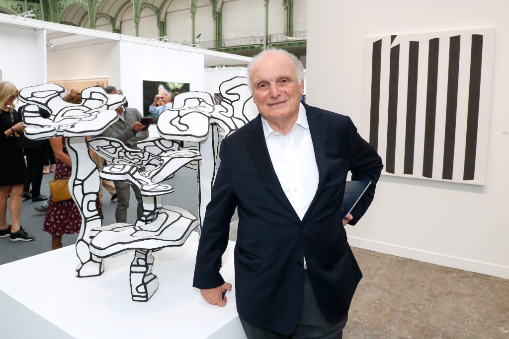 A color photo of an old, balding white man in a black suit and white shirt at an art fair with standing next to a black and white sculpture with a striped black and white painting hanging on the wall behind him, with a group of fairgoers in the background.