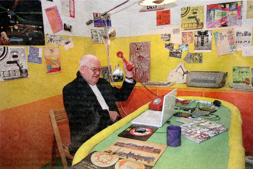 A photo of an older white man, likely in his 60s or 70s, with a bald head and glasses, seated at a brightly colored desk, holding a red telephone receiver in one hand and smiling slightly, surrounded by a collage-filled room with yellow and orange walls, vintage electronics, posters, papers, and a laptop open on the desk.