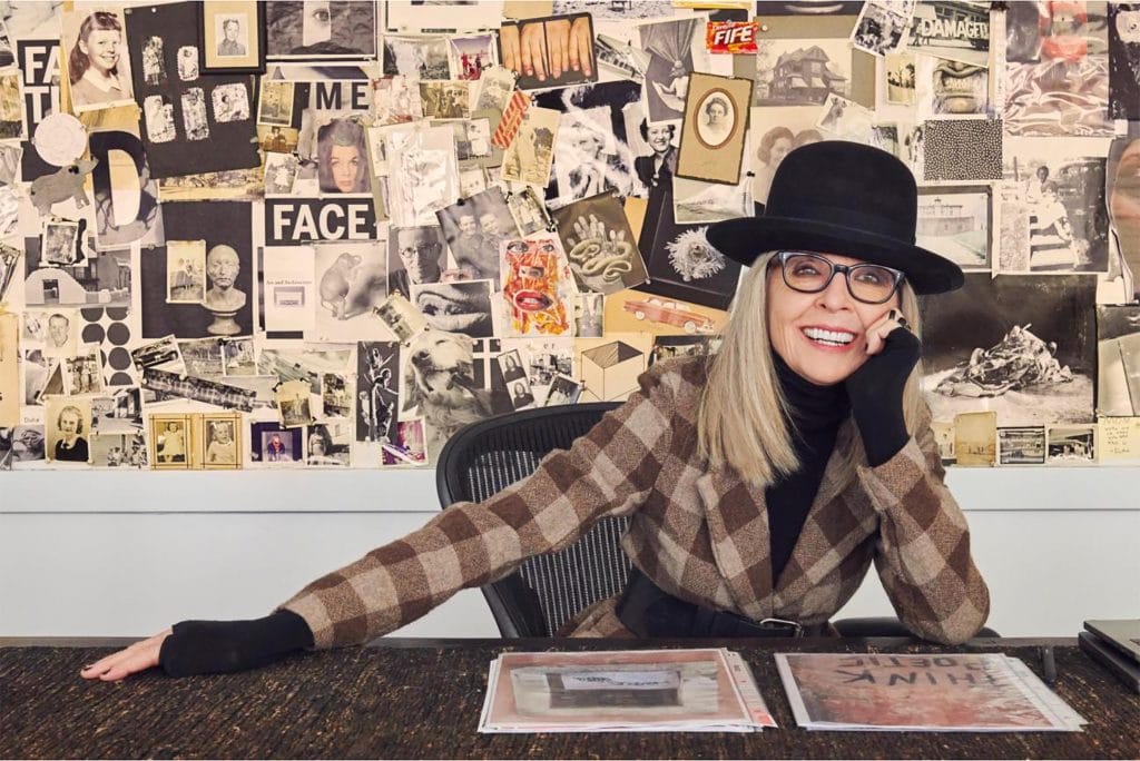 Diane Keaton posing at a desk, smiling while wearing glasses, a hat, and a plaid overcoat