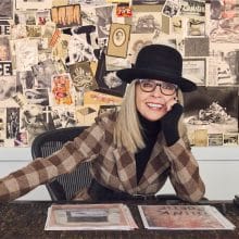 Diane Keaton posing at a desk, smiling while wearing glasses, a hat, and a plaid overcoat