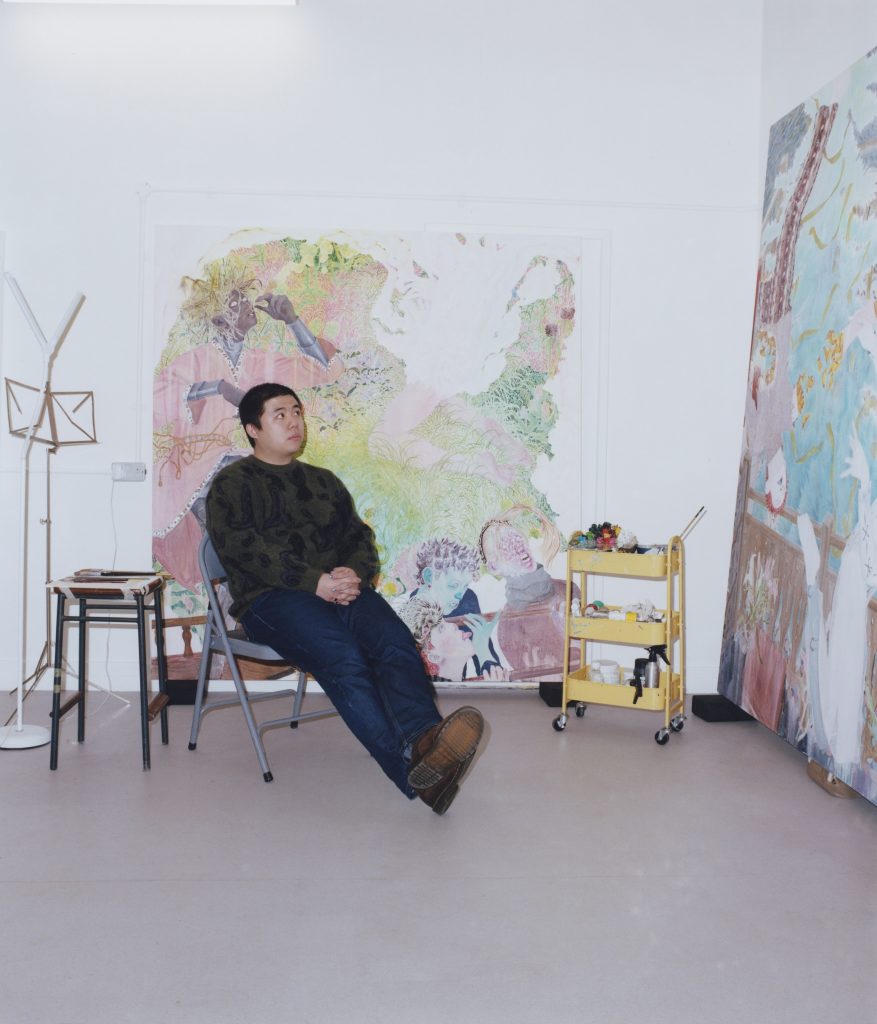 An asian man sits on a chair inside a painting-filled studio