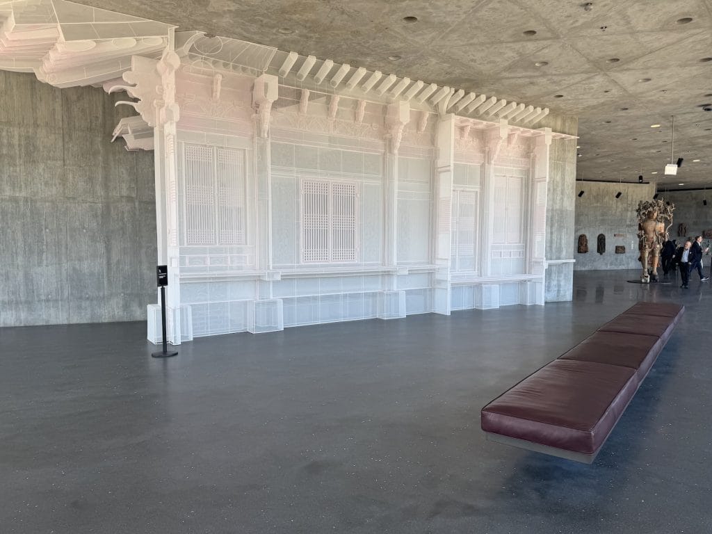 A translucent fabric architectural installation by Do Ho Suh recreates traditional Korean palace details against a concrete gallery wall, with a long burgundy bench nearby.
