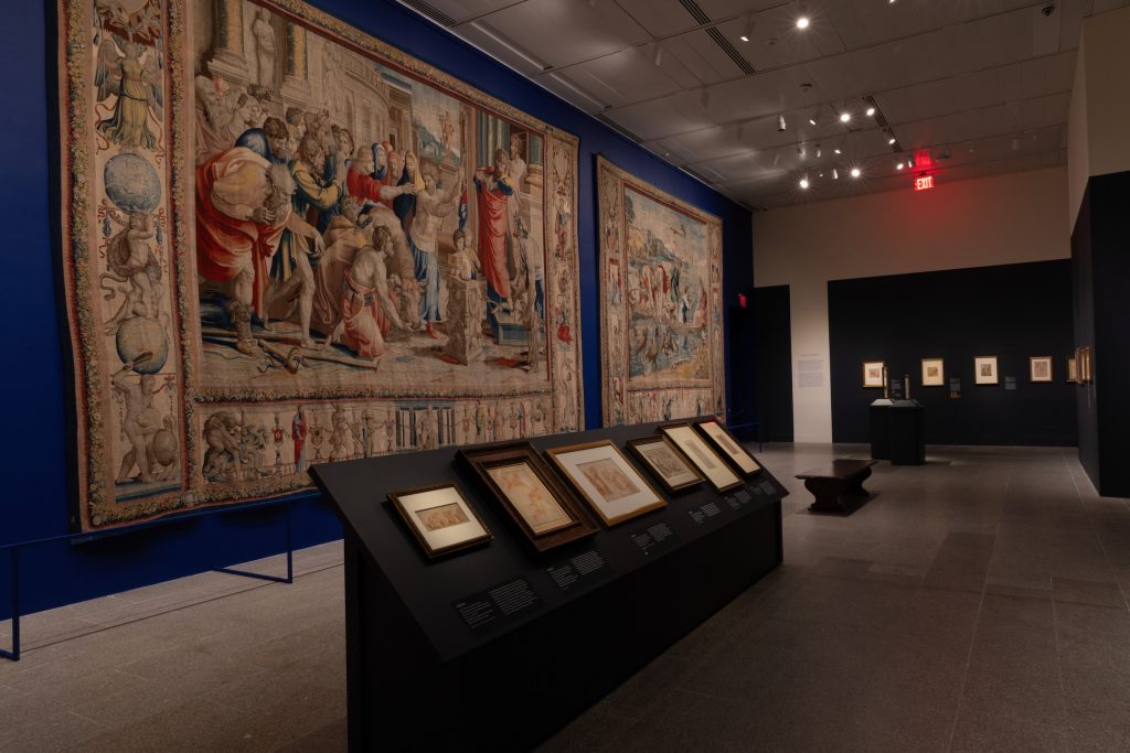 Museum gallery view featuring large Renaissance tapestries and framed drawings displayed along deep blue walls.