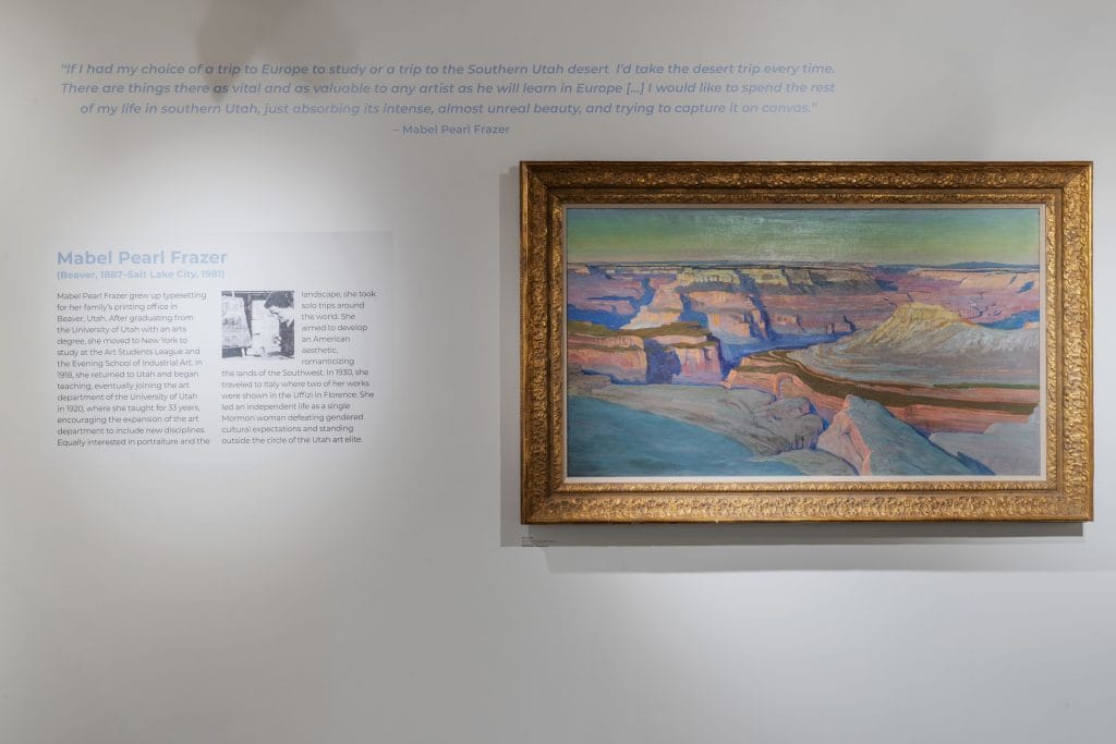 A framed landscape painting hangs on a white museum wall, depicting a colorful canyon scene with layered cliffs and a river below. To the left, a wall label features a quote and biographical text about artist Mabel Pearl Frazer, along with a small archival photograph.