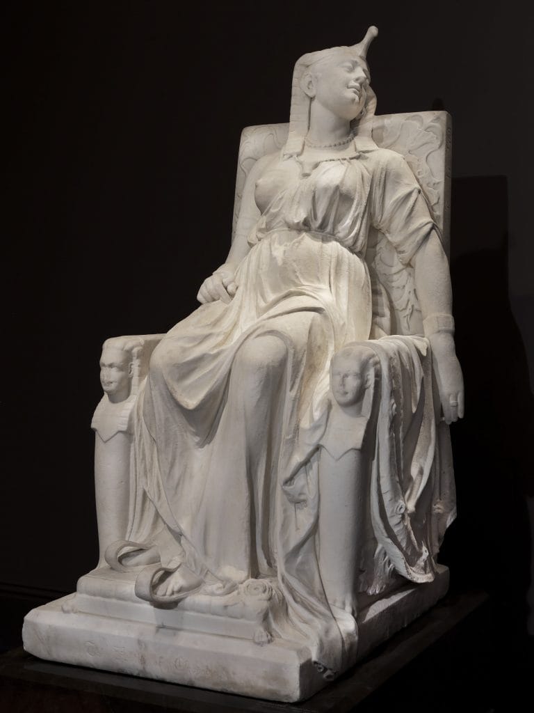A photo of a large white neoclassical marble sculpture against a black backdrop, of Cleopatra lying dead on her throne, wearing a flowing dress and Egyptian headdress.