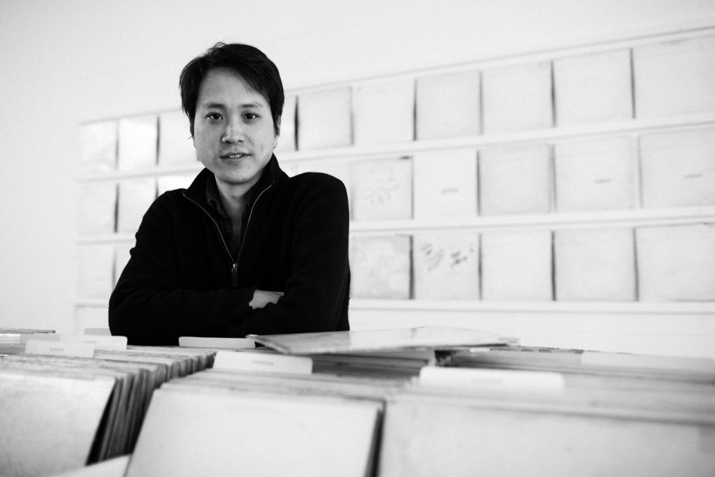 Black-and-white photograph of a person standing with arms crossed behind rows of archival boxes.