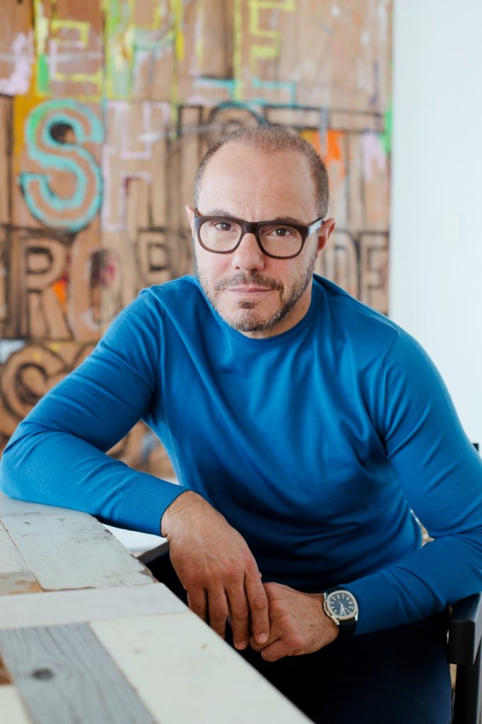 A man with short hair and glasses in a sweater poses for the camera with artworks in the background