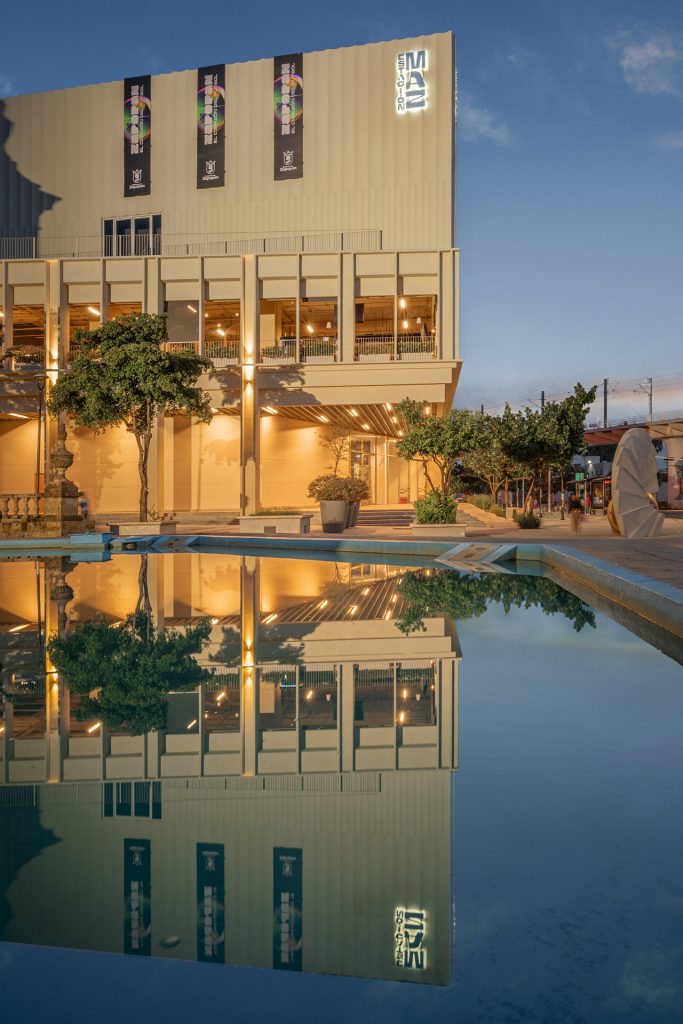 A contemporary, cream-colored cultural building at dusk with tall rectangular windows and warm interior lights glowing inside. Three vertical banners hang from the upper facade, and an illuminated sign at the top right reads “EMAZ.” In the foreground, a still reflecting pool mirrors the building, trees, and lights almost perfectly, creating a symmetrical composition. Small trees and planters line the entrance, and a large sculptural element stands to the right near a walkway and an elevated rail line in the background.