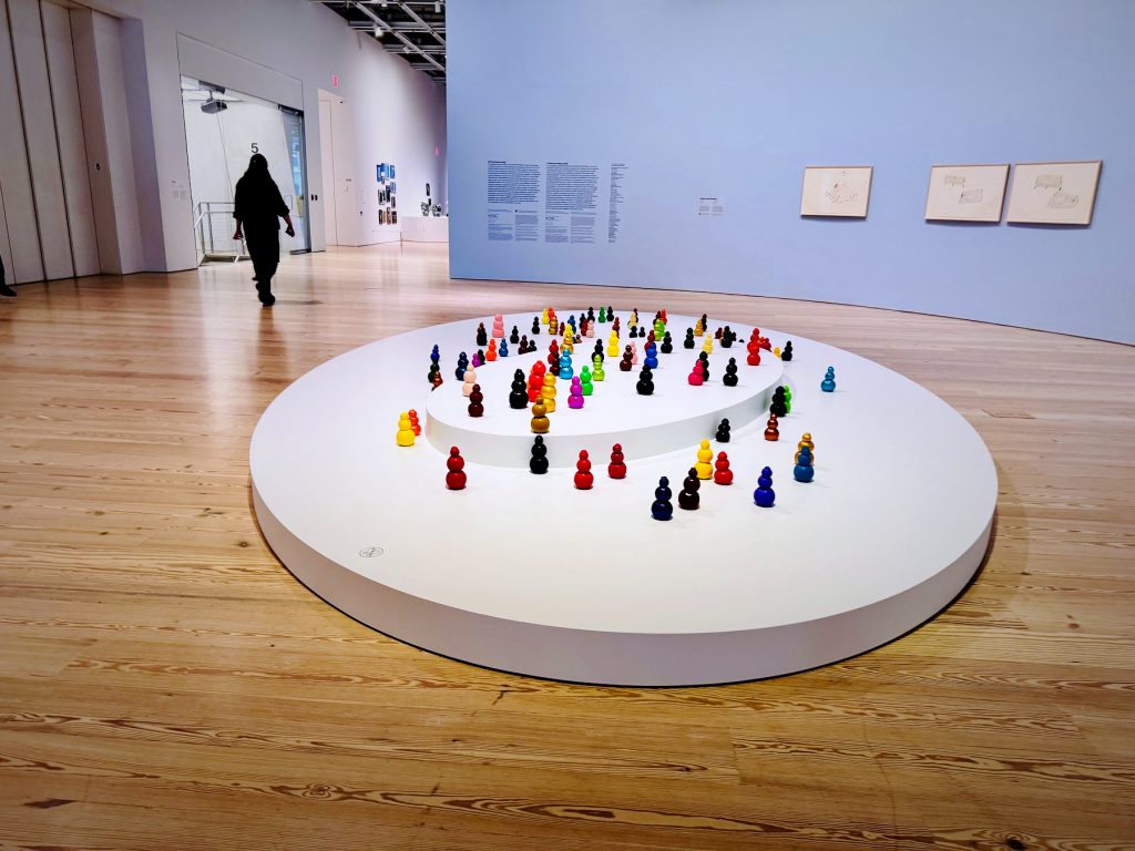 Numerous small, colorful, gourd-shaped sculptures are arranged on a tiered white circular platform in a spacious museum gallery with light wood floors and blue walls.