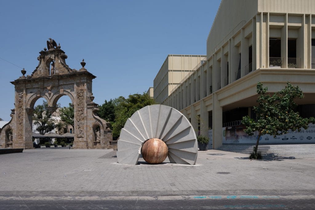 Circular sculpture with radiating concrete fins and a central stone sphere in the plaza outside Estación MAZ, next to the historic Arcos de Zapopan and a modern museum building.