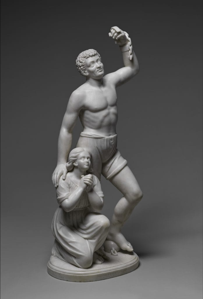 A photo of a white marble sculpture of two figures: a muscular young Black man standing with one arm raised and broken chains hanging from his wrist, and a kneeling young Black woman clasping her hands in prayer at his side, symbolizing emancipation.