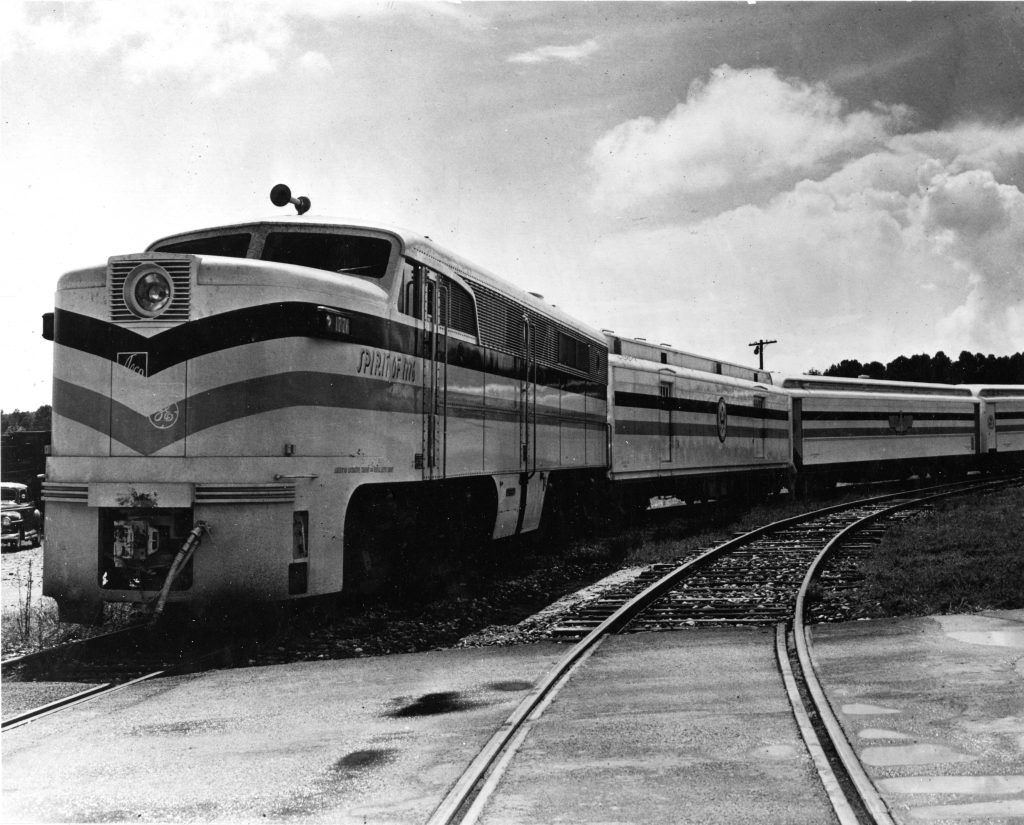 A black and white photo of the front of a large steam locomotive numbered 4449 stopped on railroad tracks, with steam drifting around the base. Tall apartment buildings and trees appear in the background, and a man stands beside a bicycle near the tracks.