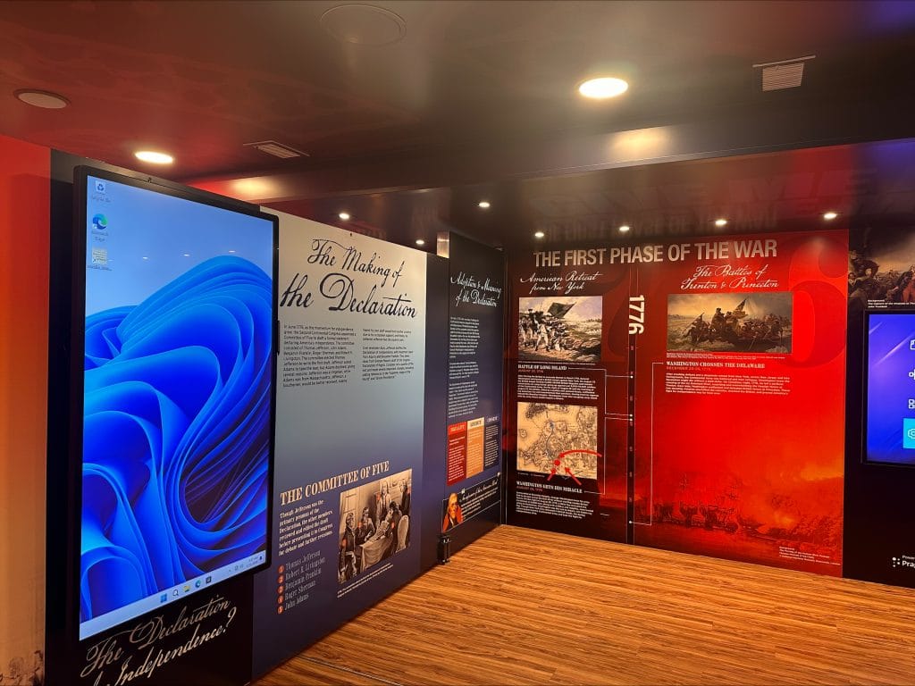 A photo of the interior of a mobile exhibit with wood flooring and large wall panels about the American Revolution. A large touchscreen monitor stands on the left beside panels titled “The Making of the Declaration” and “The First Phase of the War,” with historical images and explanatory text.
