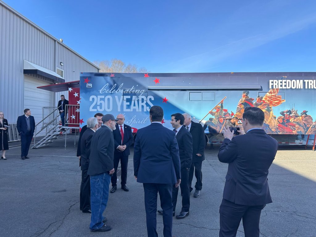 A photo of a group of adult men, mostly white and dressed in dark business suits, standing and talking in a parking lot beside a large trailer labeled “Freedom Truck” with a mural and text reading “Celebrating 250 Years of the American Spirit.” One man in the foreground holds up a phone to take a picture while others converse, with a warehouse building and a staircase leading to the truck’s entrance in the background under a clear blue sky.