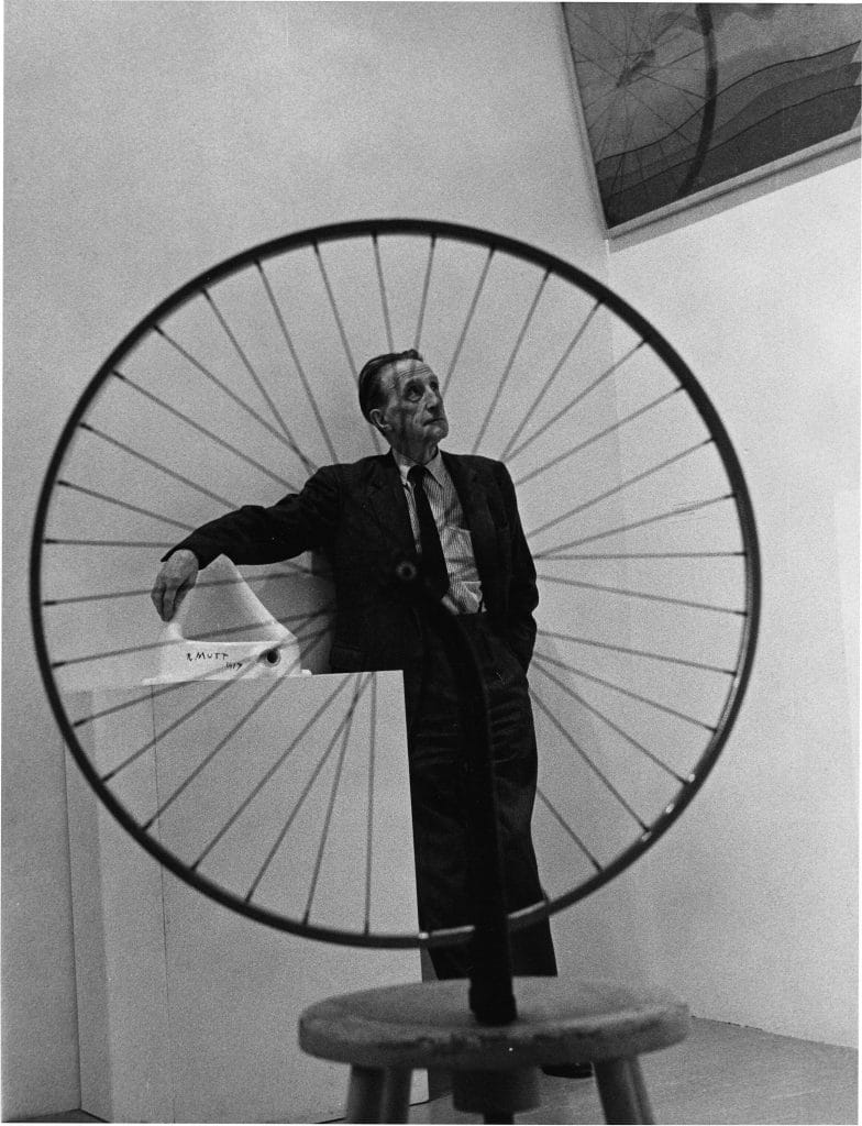 an image of the artist standing behind his bicyle wheel sculpture