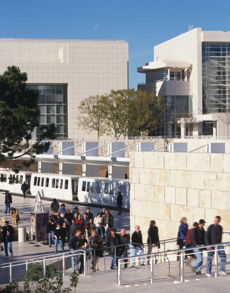 A photograph of a white tram at a passenger platform amidst a pristine white museum campus, beneath bright blue skies