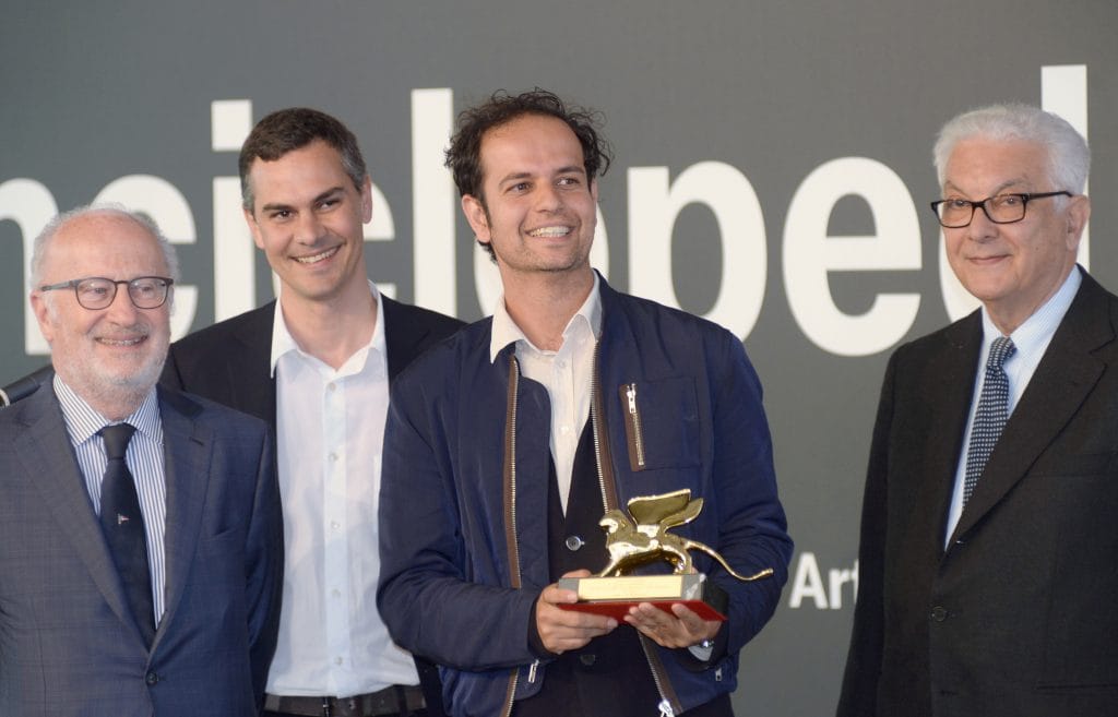 Tino Sehgal smiles while holding the Golden Lion trophy, flanked by Massimiliano Gioni and Paolo Baratta at the Venice Biennale.