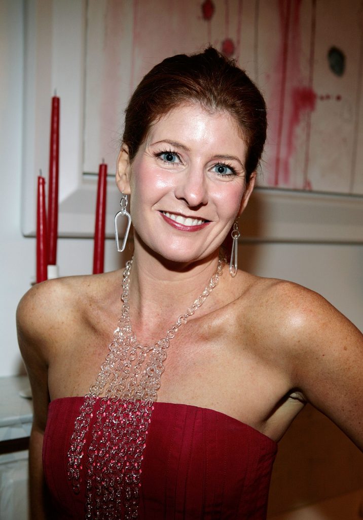 Smiling woman at an indoor event wearing a strapless red dress, long chain-style necklace, and dangling earrings, standing in front of abstract artwork and red candles.