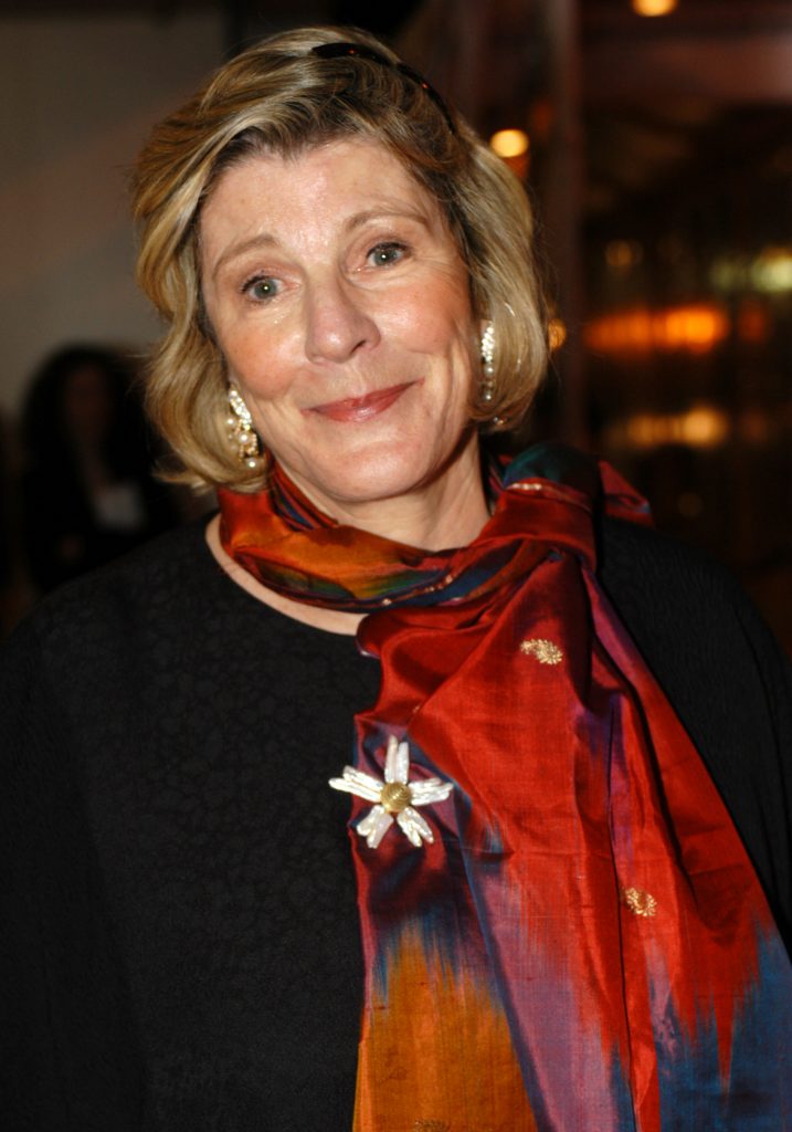 Elegant older woman with short blonde hair wearing a colorful silk scarf and pearl earrings, smiling at an indoor event.