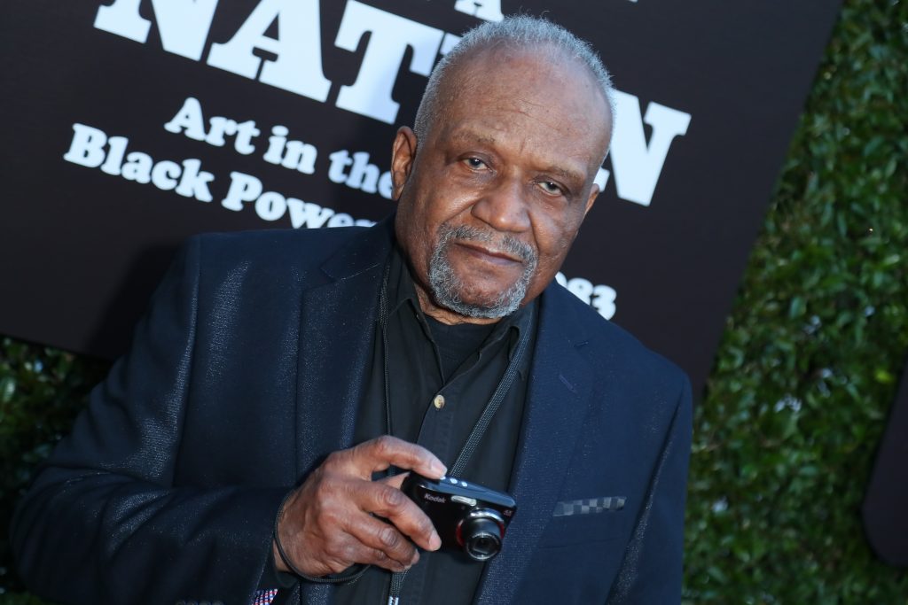 Man holding a camera while posing at an art exhibition event, standing in front of signage related to Black Power–era art, in a polished documentary-style portrait.