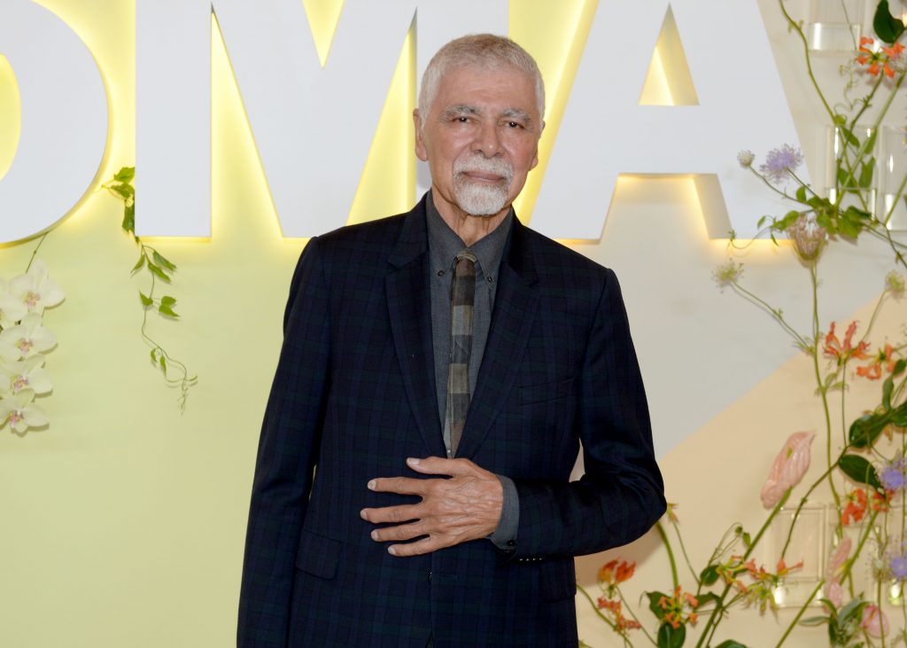 An older man with short gray hair and a goatee stands in front of a decorative backdrop with large white letters and flowers, wearing a dark suit and patterned tie.