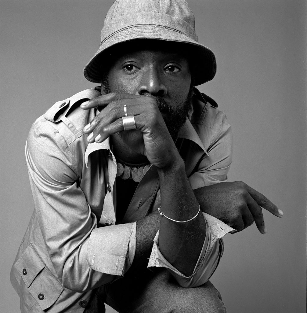 Black-and-white studio portrait of a man in a bucket hat and casual jacket, captured in a thoughtful pose that highlights expressive gesture and classic portrait photography style.