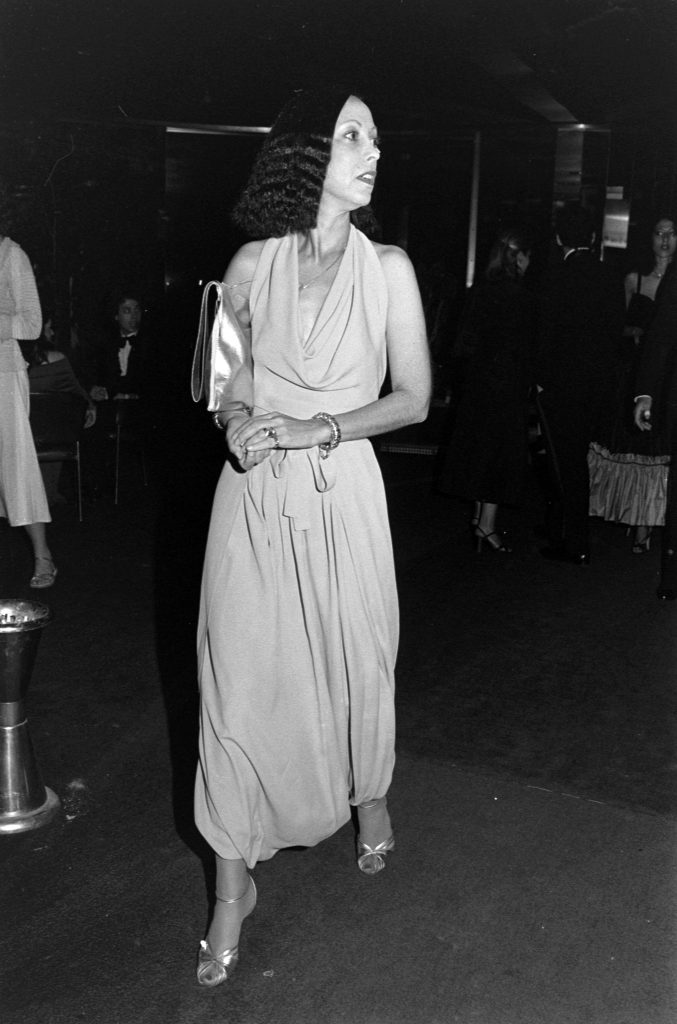 Elegant woman in a draped evening gown walking through a dimly lit event space, holding a clutch and wearing heeled sandals.
