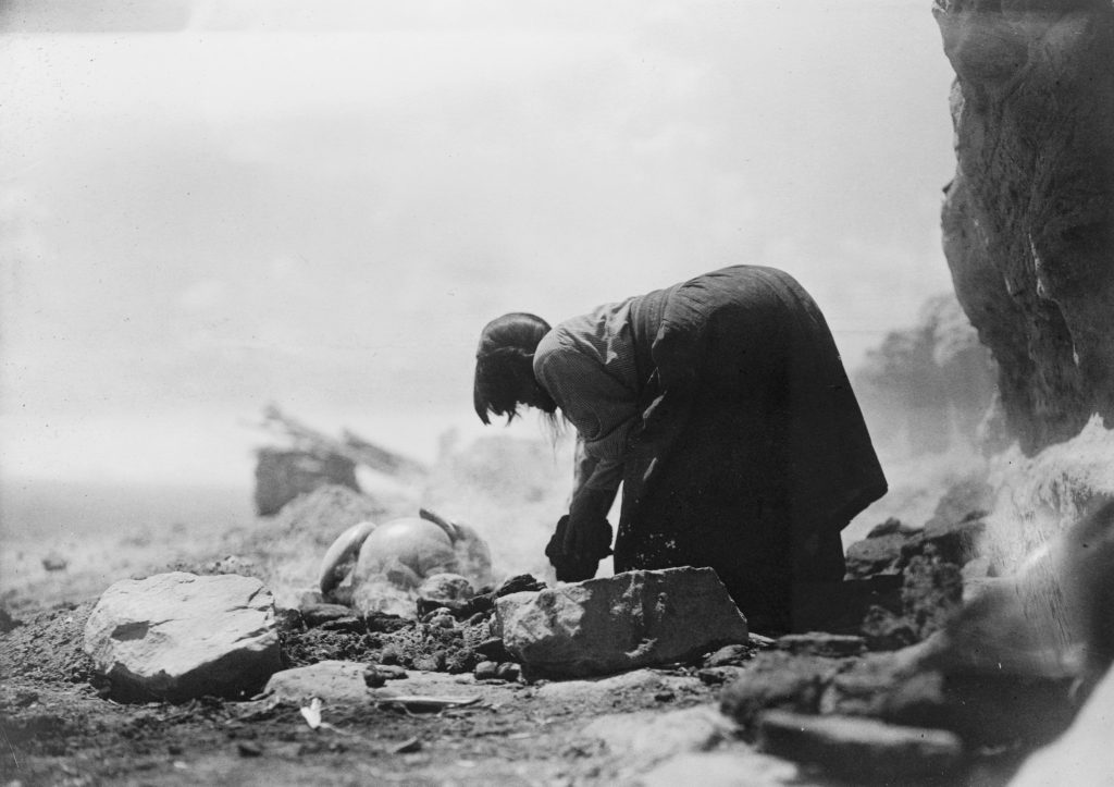 Black-and-white photograph of a person crouched on the ground shaping pottery by hand outdoors, surrounded by rocks and rough terrain; a finished pot lies nearby.