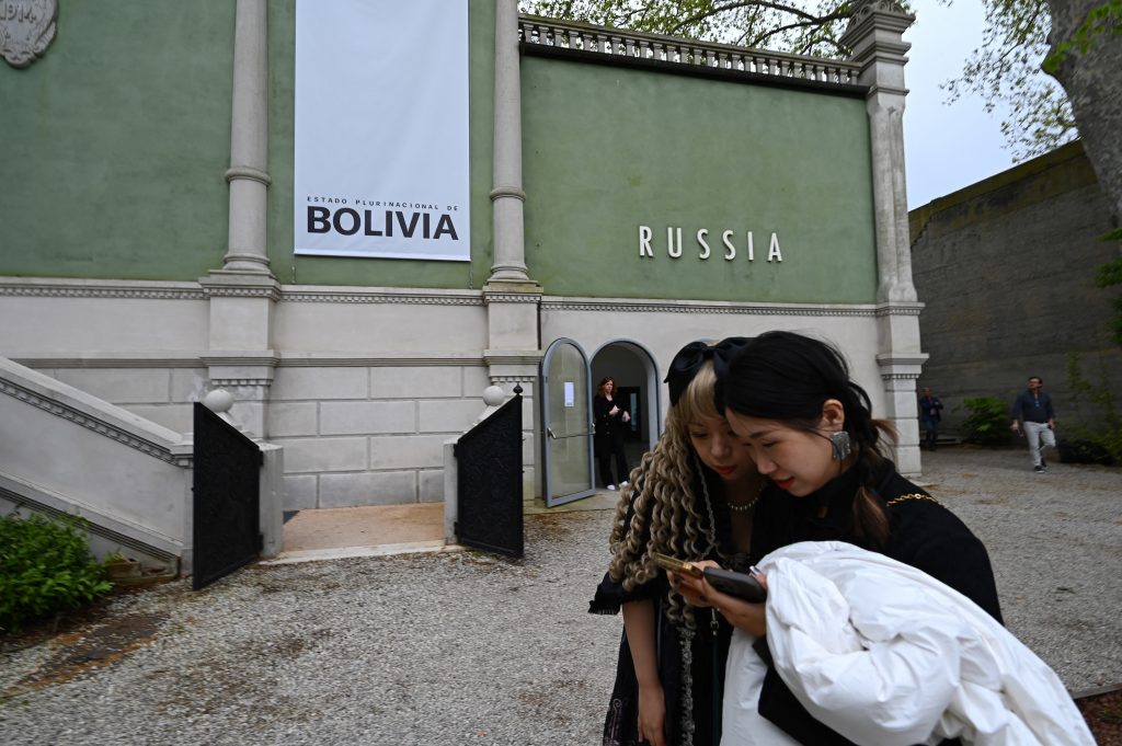 Two women look at a smartphone in front of a pavilion marked "Russia" and "Bolivia" at the Venice Biennale