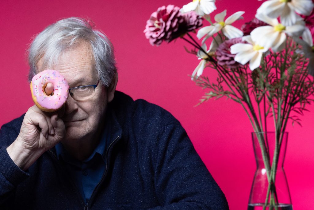 Older man playfully holding a pink frosted donut over one eye beside a vase of flowers against a bright magenta background.