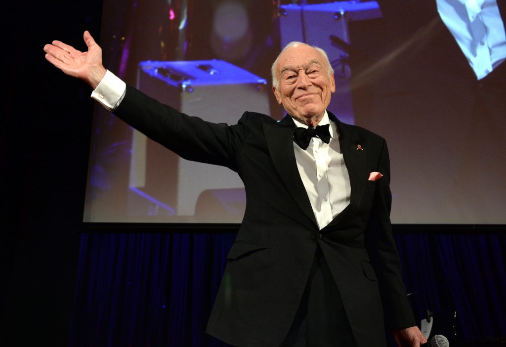 Elderly man in a black tuxedo smiling and raising one arm on stage at a formal event, standing before a large screen projection behind him.