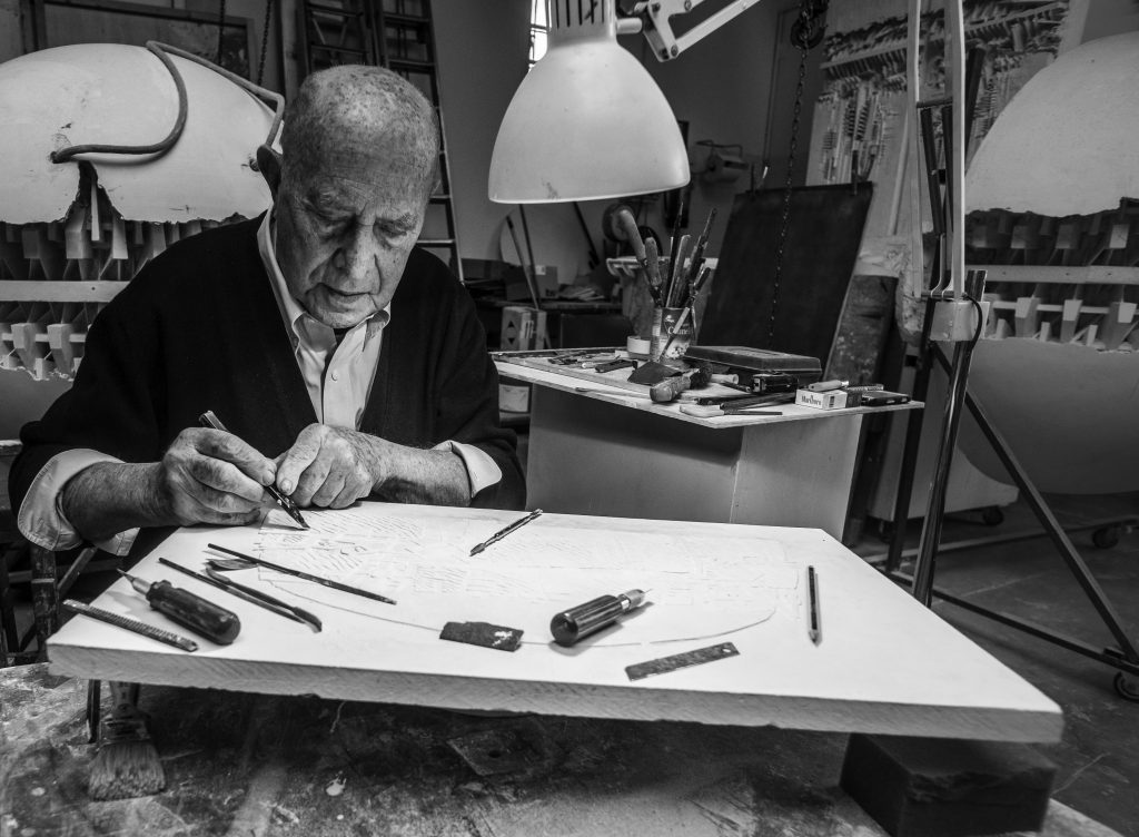 Artist carving a detailed relief in a studio surrounded by sculpting tools, shown in a black-and-white photograph emphasizing craftsmanship and the creative process.