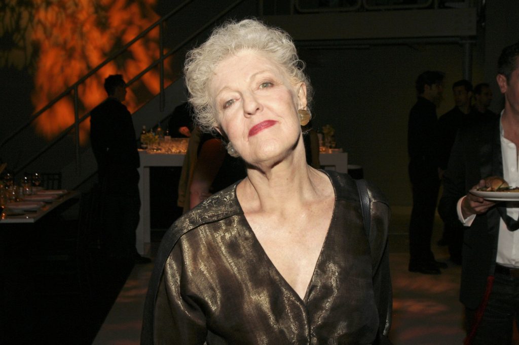 Elegant older woman with short curly hair and red lipstick posing at an evening event, illuminated by warm ambient lighting with guests mingling in the background.