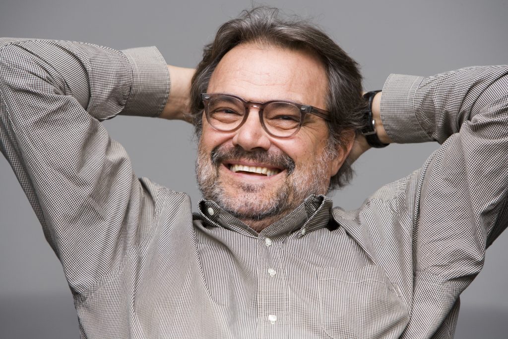 Smiling man in glasses leaning back with hands behind his head in a relaxed studio portrait, highlighting expressive personality and contemporary editorial photography style.