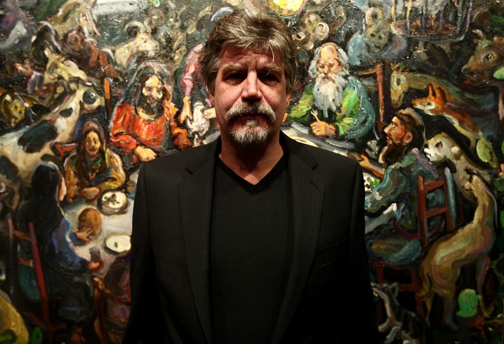 Man in a black suit standing in front of a vibrant, expressive painting depicting a crowded table scene with richly textured, colorful figures.