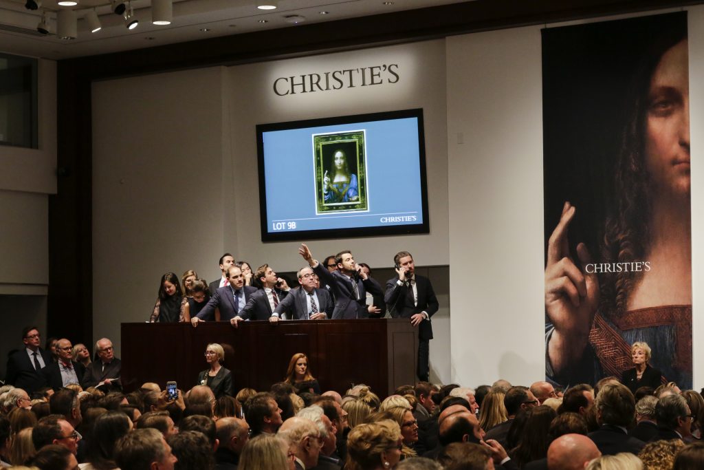 People in a crowded room raise their arms, competing for an artwork