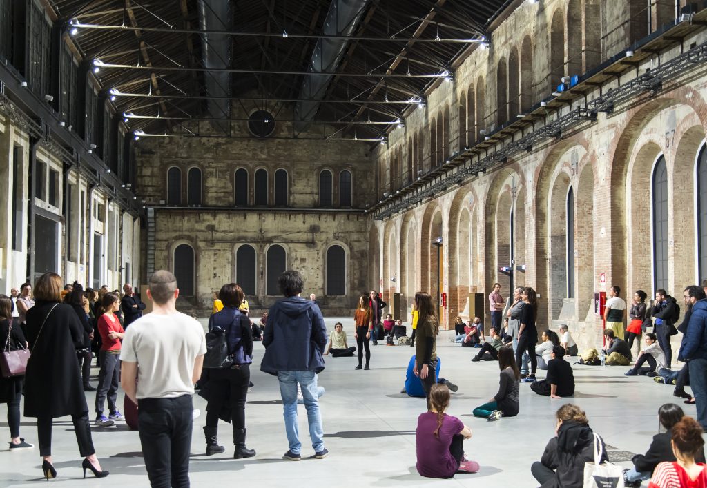 A large group of people gather inside a vast industrial hall with high wooden beams and tall arched brick walls. Some stand in a wide circle while others sit on the floor, facing inward toward a few individuals in the center. The space resembles a renovated warehouse or contemporary art venue, with exposed brick, tall windows, and overhead lighting illuminating the open concrete floor.