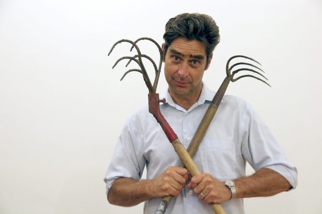 Man in a light button-down shirt holding two rustic metal garden tools crossed in front of him against a plain white background.