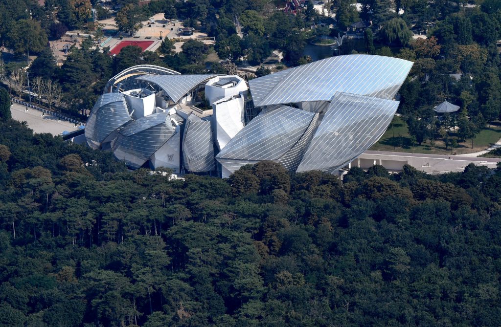 An aerial view of a large, modern building composed of multiple curved, glass-paneled structures, surrounded by dense trees in a park setting.