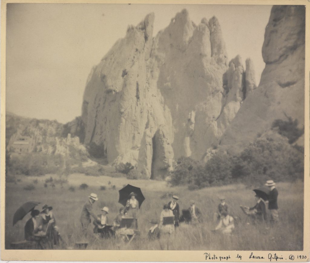 A sepia toned photograph of men and women painting and sketching on the grass with huge mountains looming above them