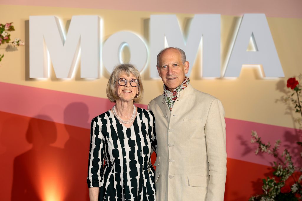 A photo of an older woman and man standing together at a MoMA event. The woman, with short blonde hair and glasses, wears a black-and-white patterned dress with a pearl necklace, while the man, with short gray hair, wears a light beige jacket with a floral scarf at his collar. They are smiling in front of a colorful wall with large white “MoMA” letters and floral decorations.