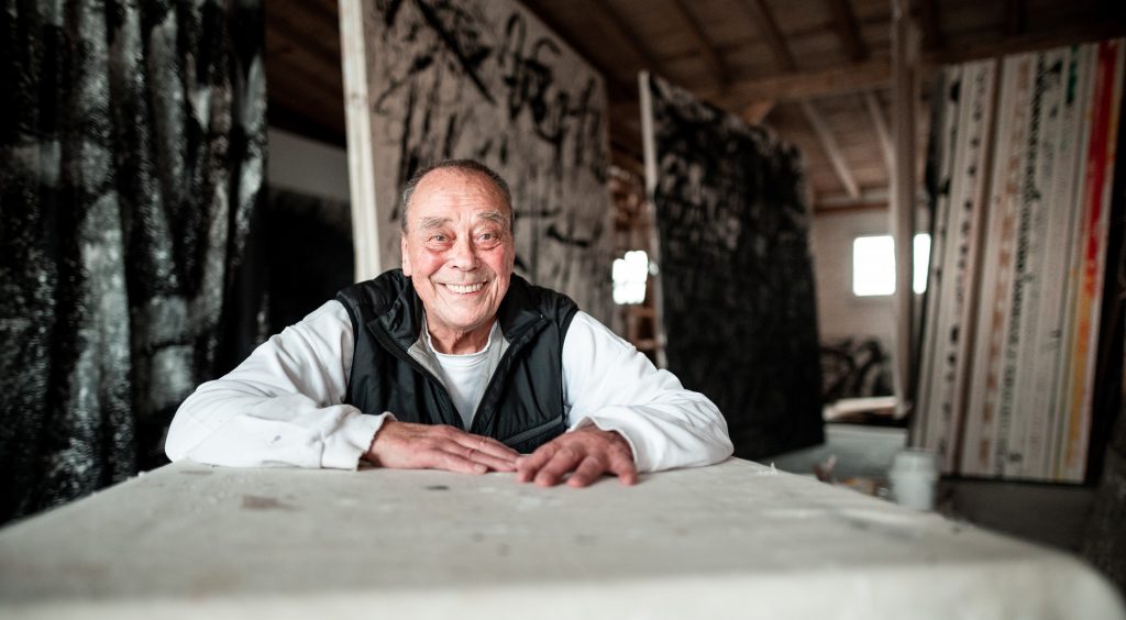 Smiling older man leans on table, surrounded by abstract monochrome paintings in rustic-lit studio